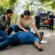 A bystander gives first aid to a collapsed person while someone calls emergency services and paramedics arrive.